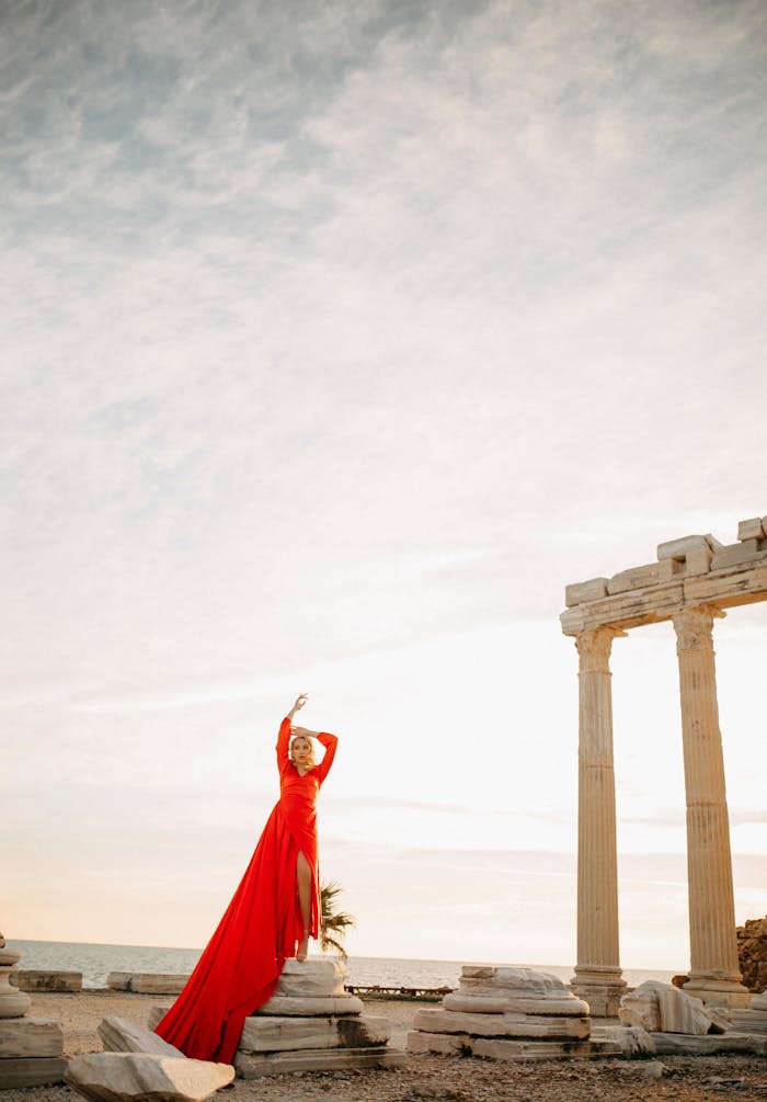 A striking woman in red dress poses elegantly amid ancient pillars, under the wide open sky.