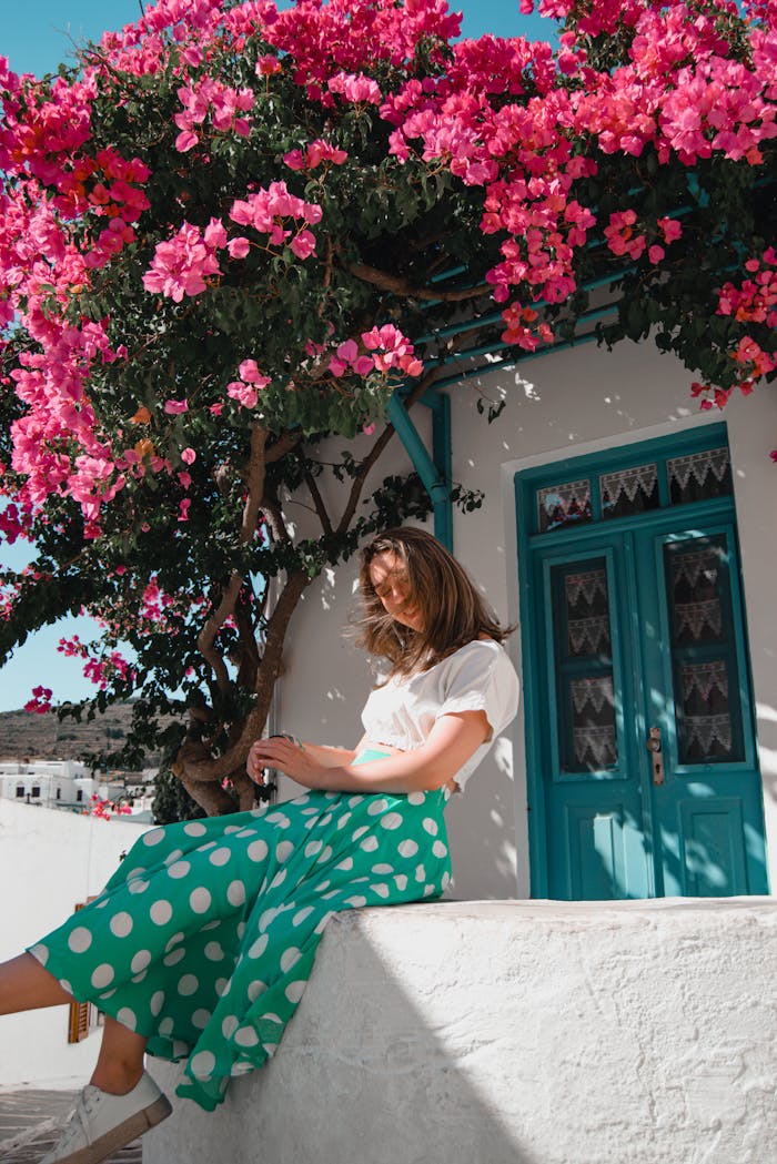 A woman enjoys a sunny day wearing a white blouse and green polka dot skirt near vibrant pink flowers.
