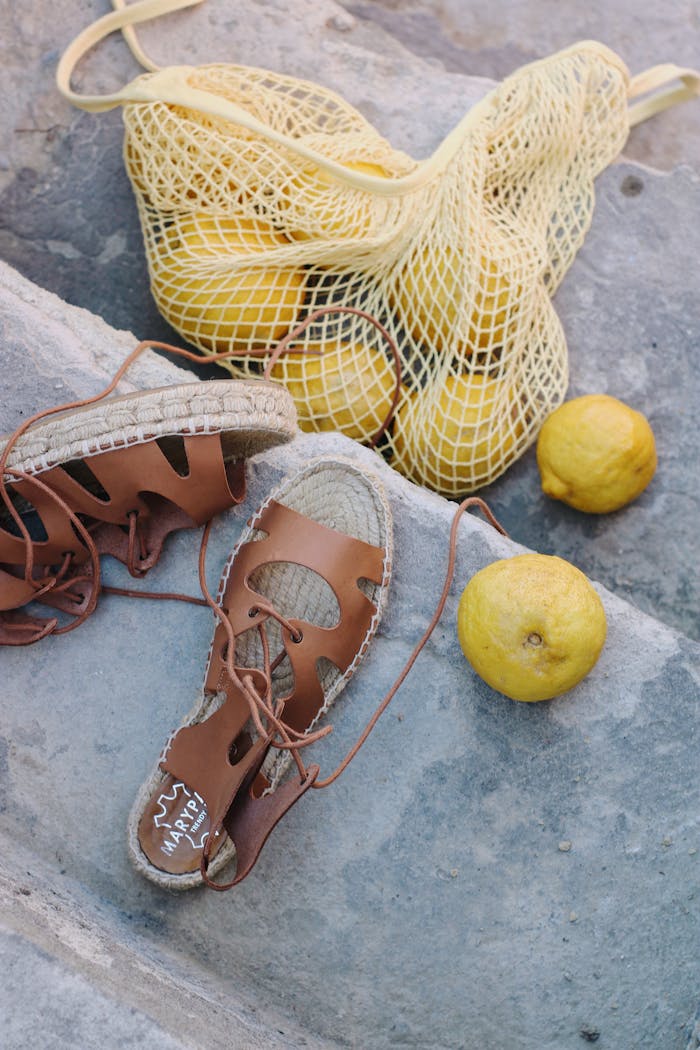 Brown sandals next to a mesh bag of fresh lemons on a stone surface, summer vibes.